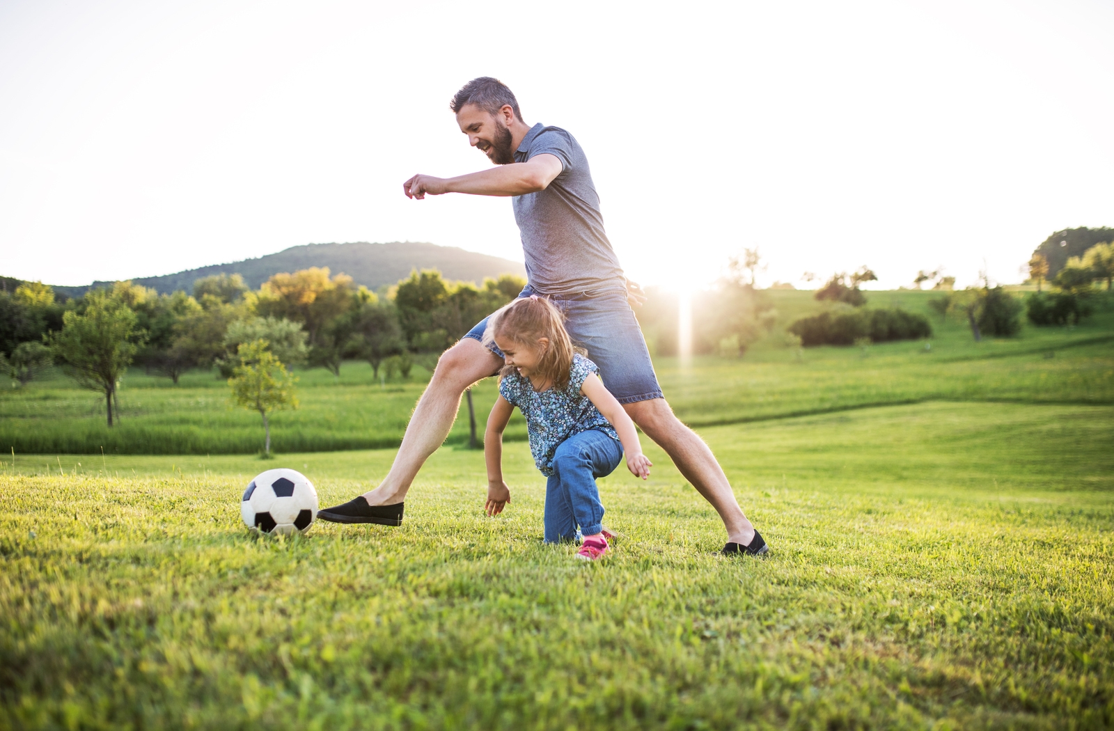 A dad and his young daughter play with a soccer ball in a green field with trees behind them.
