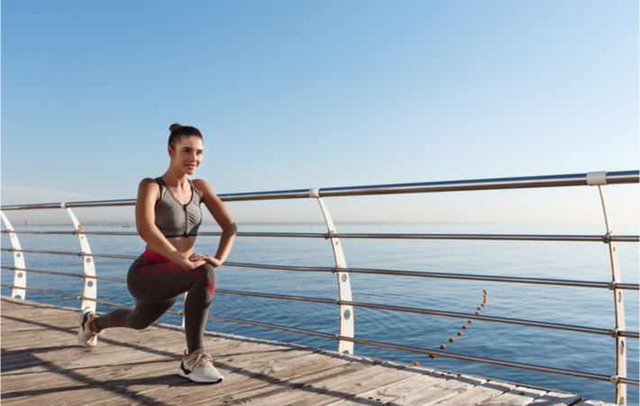 A woman wearing workout clothing does lunges along a wooden promenade next to a waterbody.