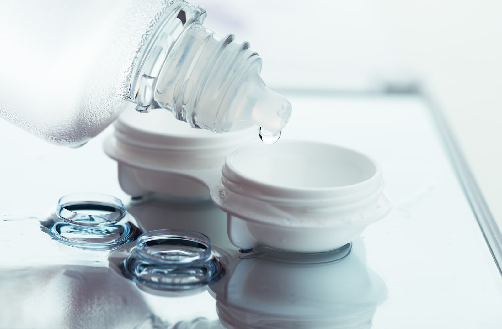 A pair of contact lenses sitting next to a contact lens case and someone pouring contact lens solution in the case
