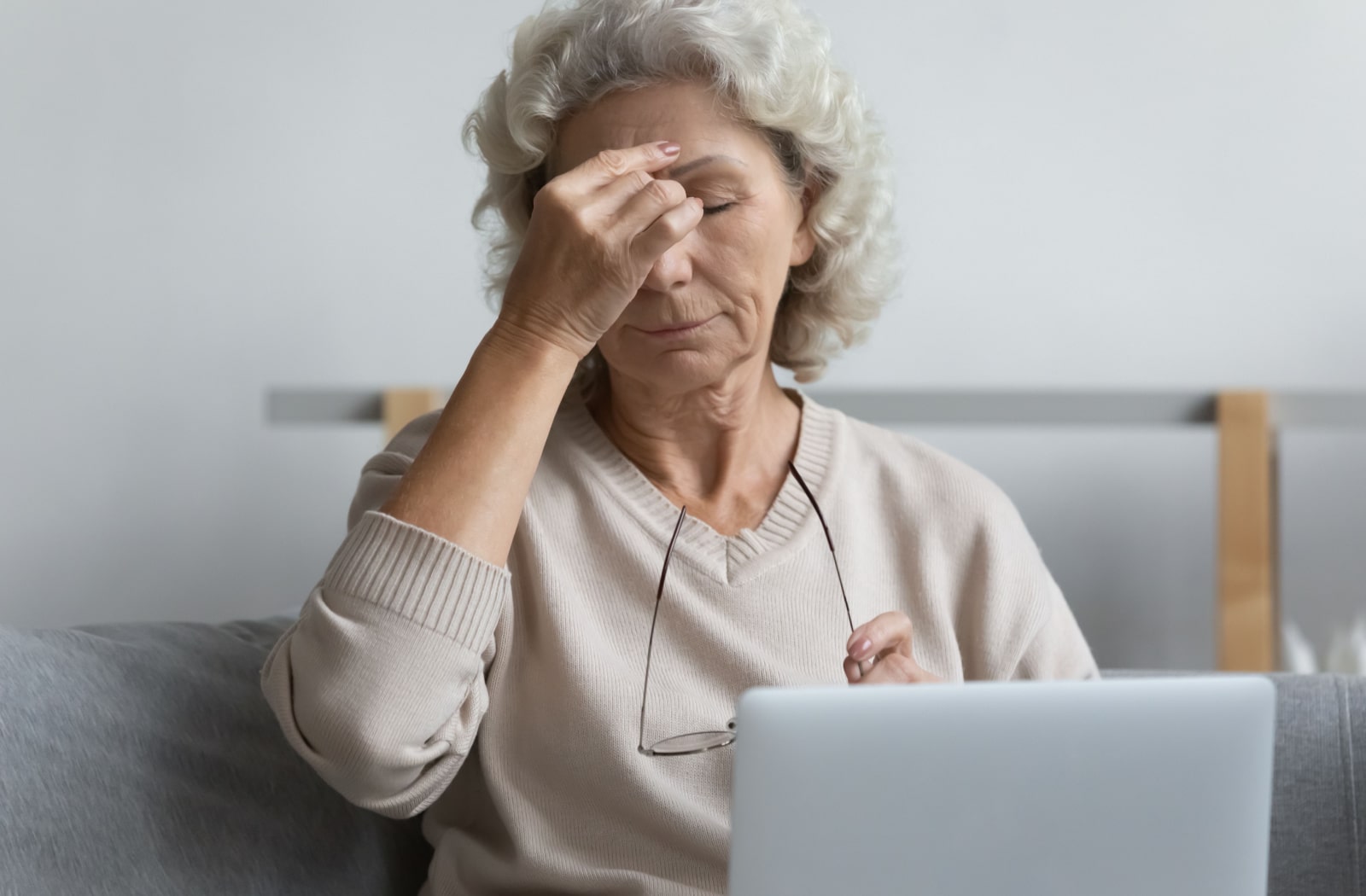 A middle-aged woman sitting on a gray couch with a laptop sitting on her lap. She is taking off her glasses and rubbing the bridge of her nose due to eye pain.