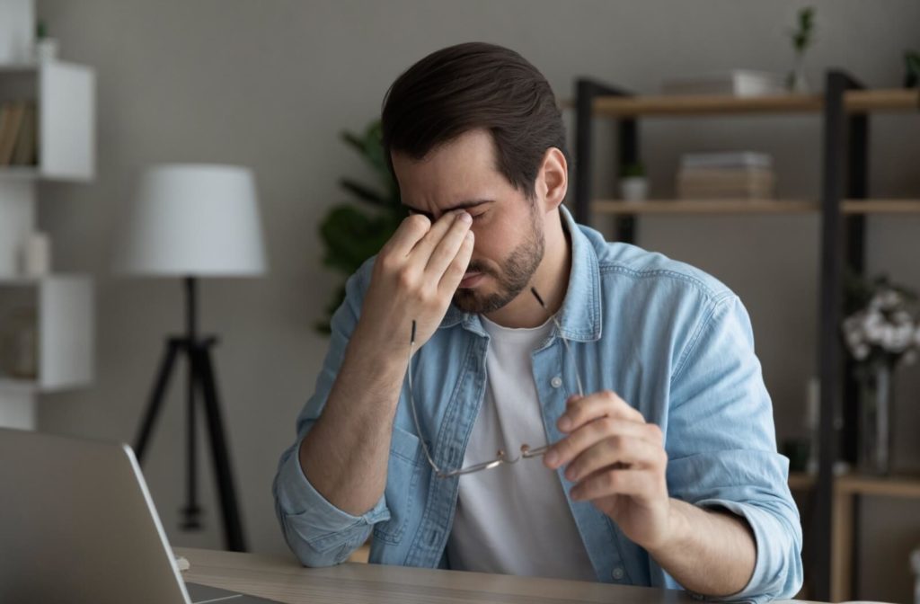 A patient sitting in front of a laptop at a desk holds their glasses in one hand while rubbing their itchy eyes with the other.