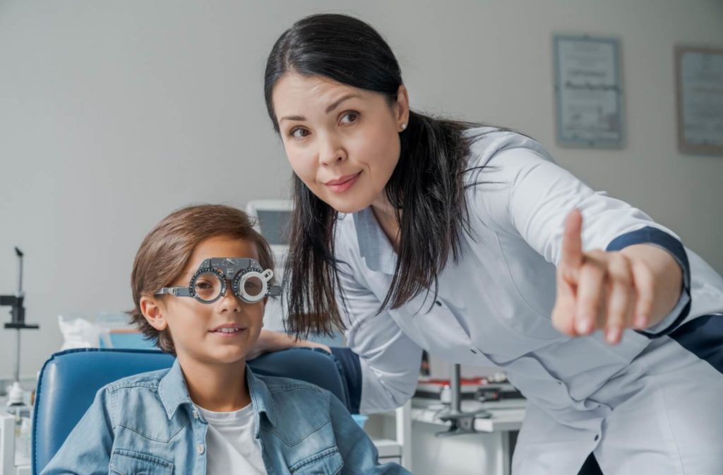 child is sitting in an eye exam chair with a phoropter (eye exam device) on, while an optometrist points and provides instructions.