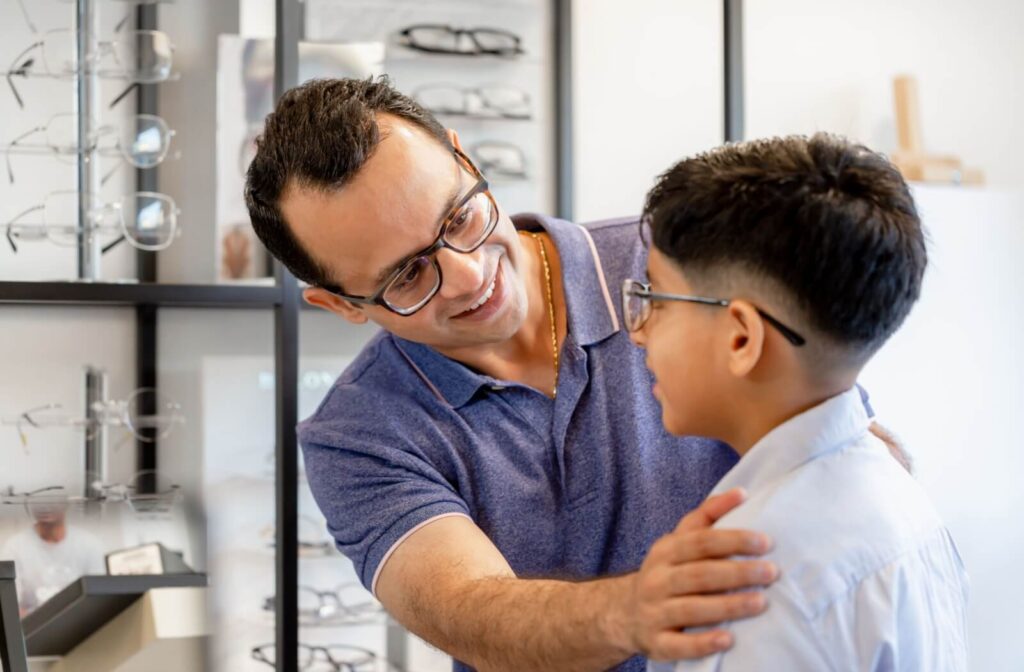 A father adjusts his son's glasses while they smile at each other inside an optometry clinic.