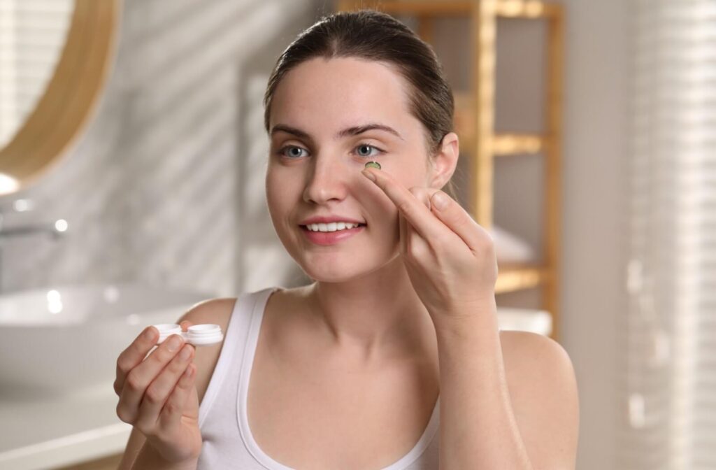 Smiling woman applying a coloured contact lens in front of a bathroom mirror while holding a lens case.