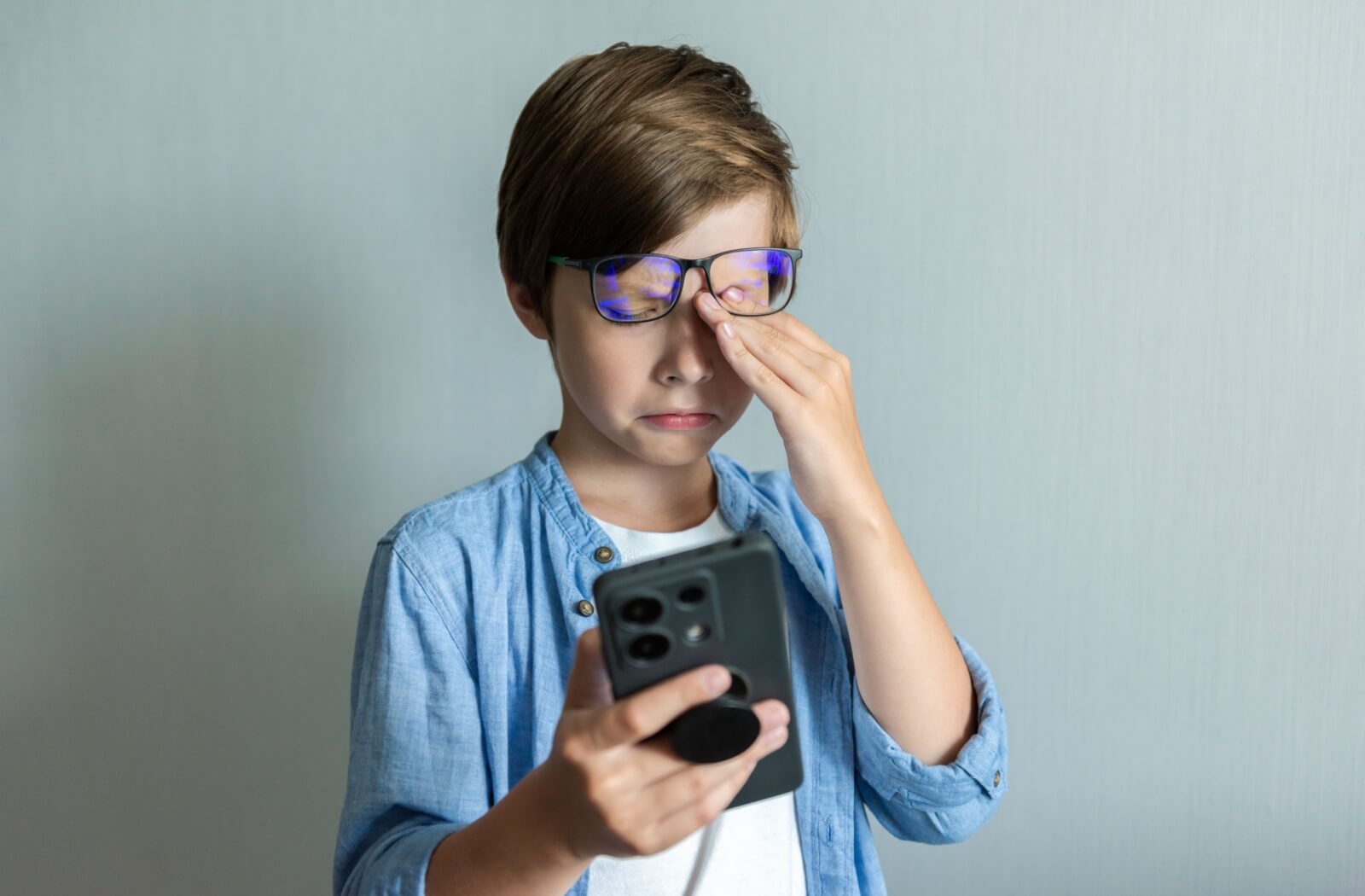 Young boy wearing blue-light glasses rubbing his eye due to digital eye strain while using a smartphone.