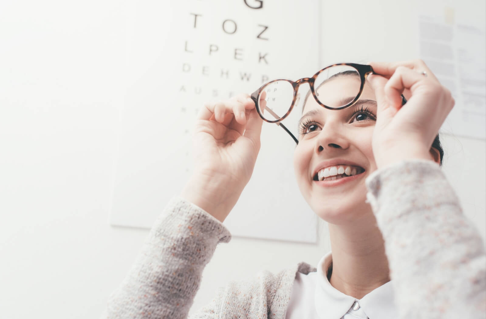 Young woman smiling as she tries on new glasses in an optometrist’s office with an eye chart in the background.