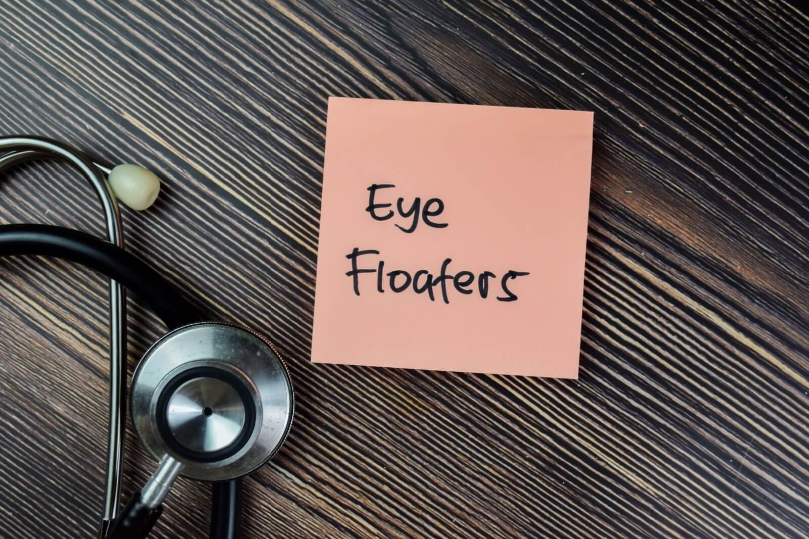 A pink sticky note with the handwritten words "Eye Floaters" placed on a dark wood surface next to a medical stethoscope.